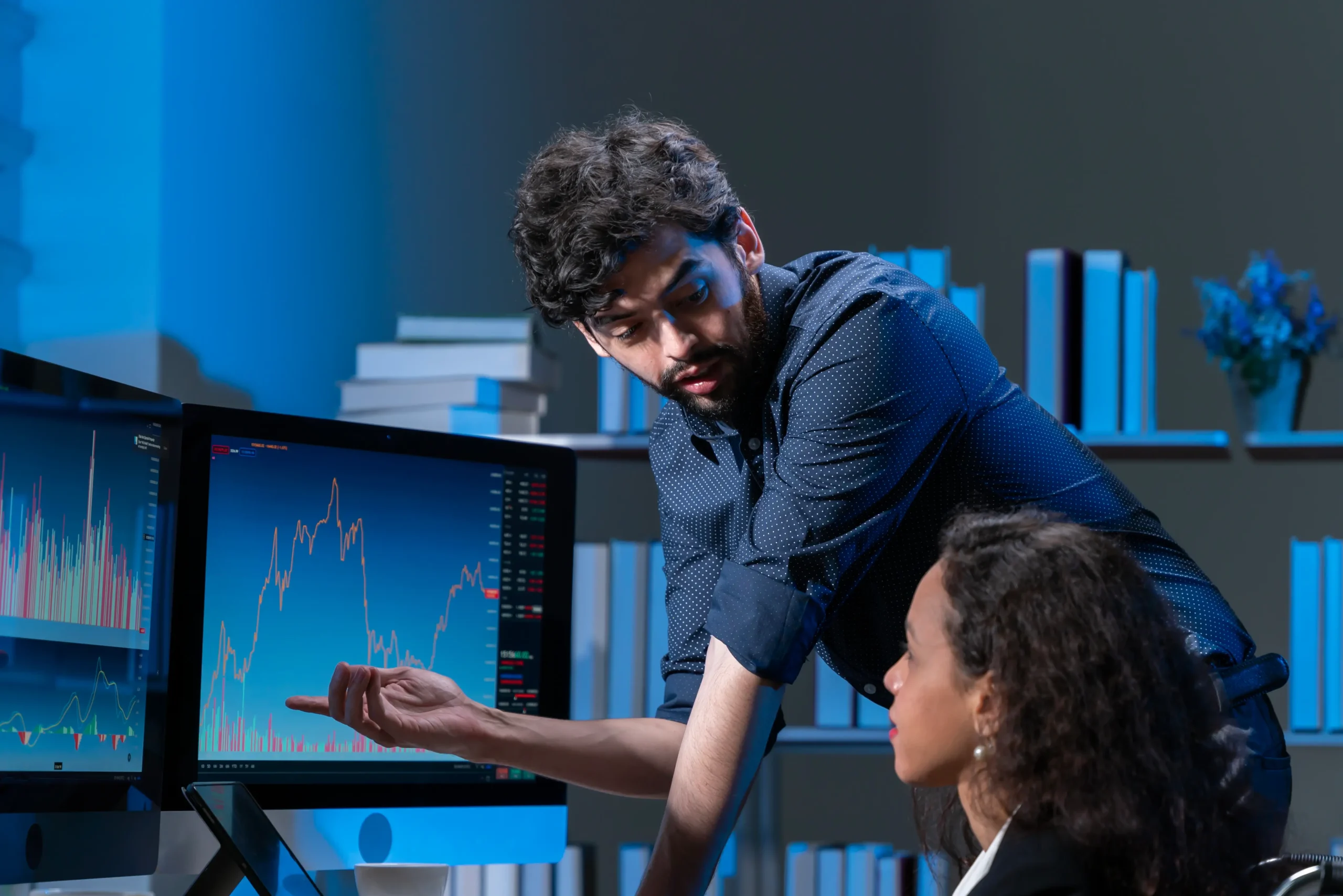 A man explains financial charts on a computer monitor to a woman in an office with shelves of books in the background.