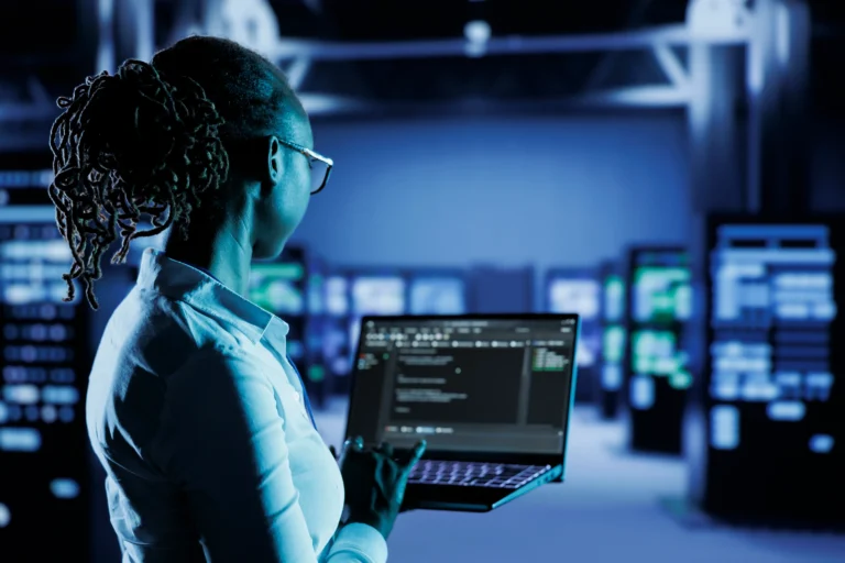 Person holding a laptop with code on the screen, standing in a server room with rows of servers and blue lighting in the background.