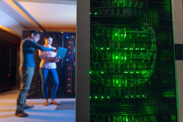 Two people stand in a server room discussing data on a laptop, with server racks and green indicator lights visible in the foreground.