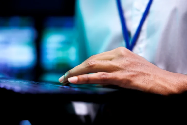 A close-up of a person's hand using a touchscreen device, with the individual wearing a white shirt and a blue lanyard.