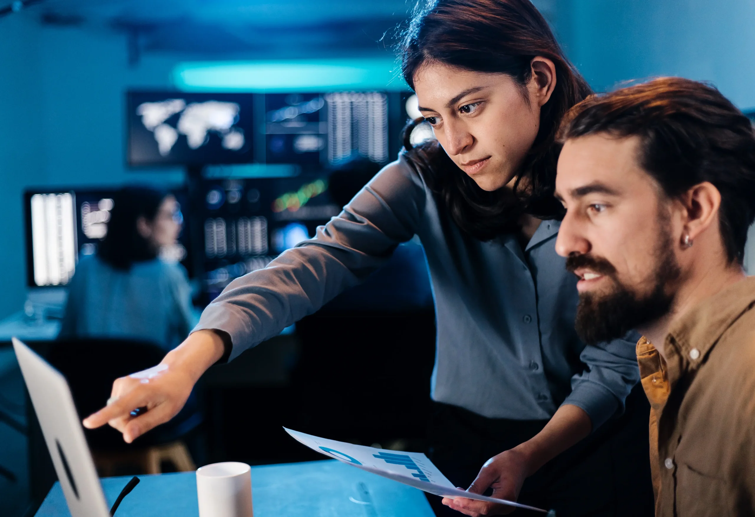 Two people look at a computer screen in an office with data charts; one points at the monitor while holding a document, with coworkers visible in the background.