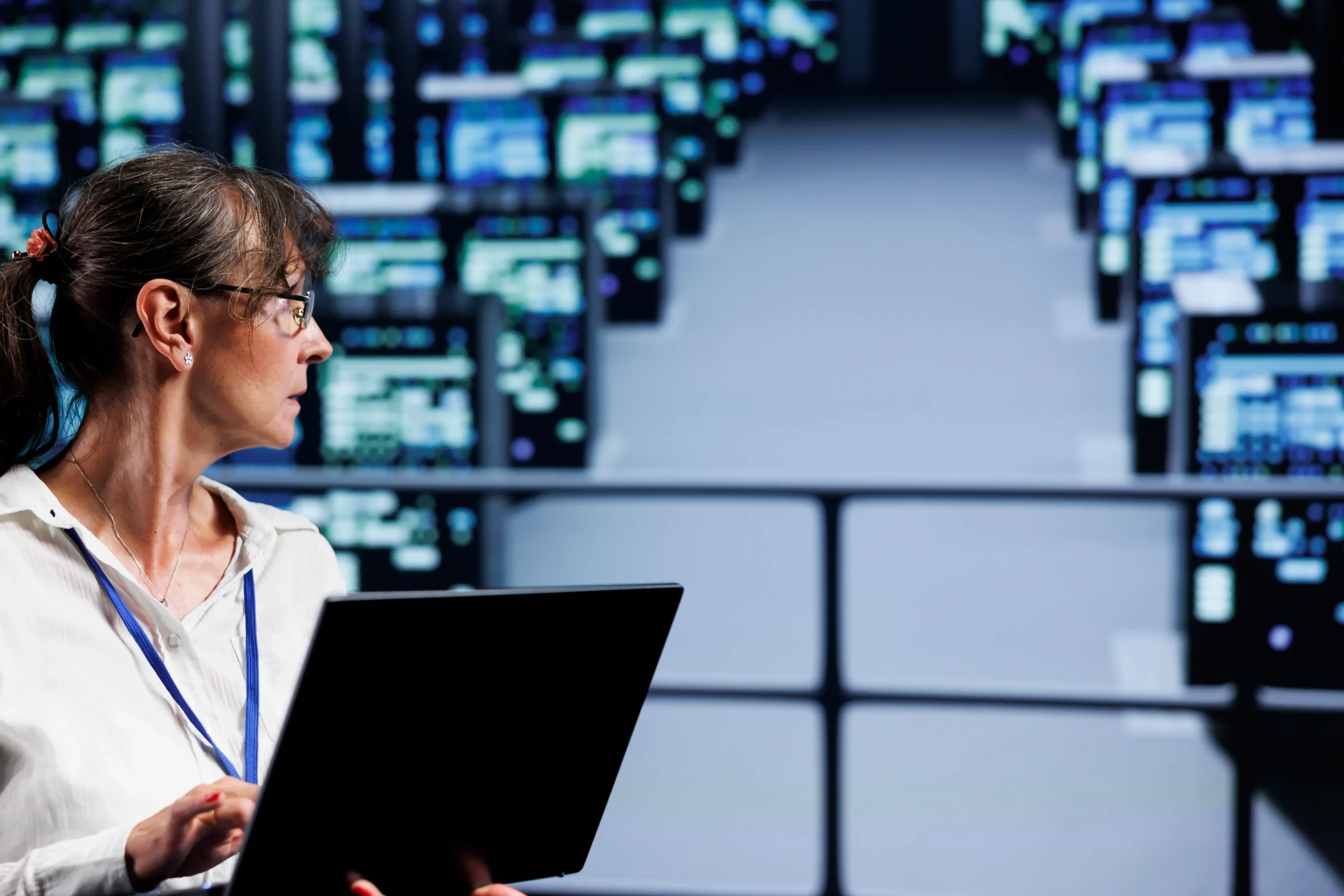 A woman holding a laptop stands in front of multiple computer monitors displaying data in a control or server room environment.