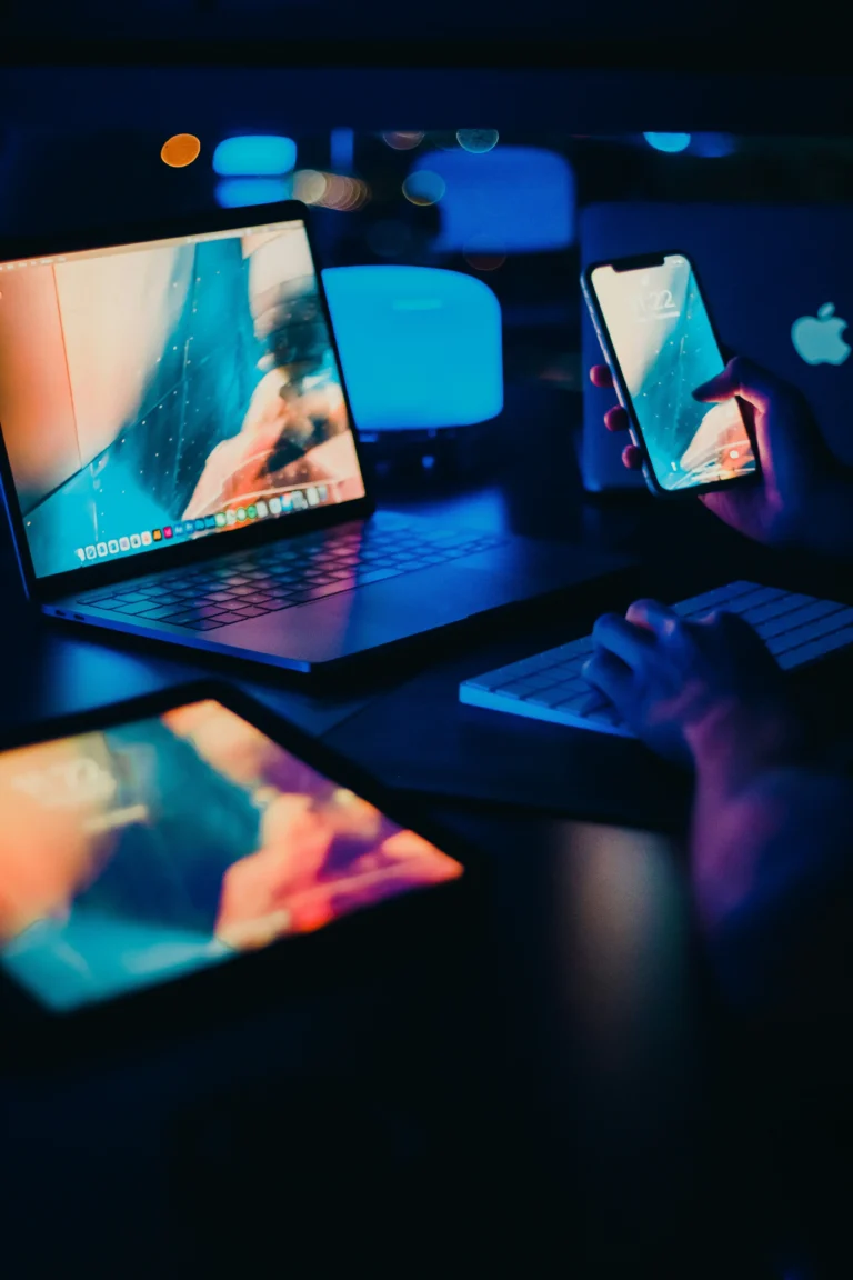 A person using a smartphone, with a laptop, tablet, and another laptop on a desk, all displaying similar blue-toned screens in a dark room.