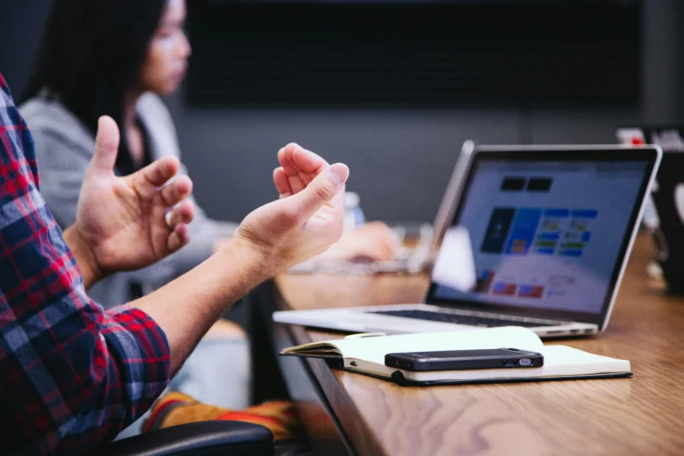 Two people sit at a desk with a laptop, notebook, and phone; one person gestures with their hands during a discussion or meeting.