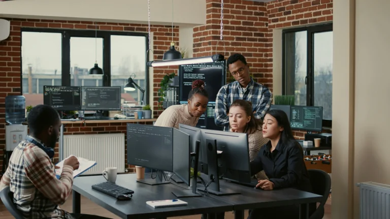 Four people gather around computer monitors in a modern office, discussing code displayed on the screens. Another person sits nearby with a clipboard. Brick walls and large windows are visible.