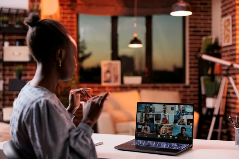 A woman sits at a desk in a home office, participating in a video conference call with multiple people displayed on her laptop screen.