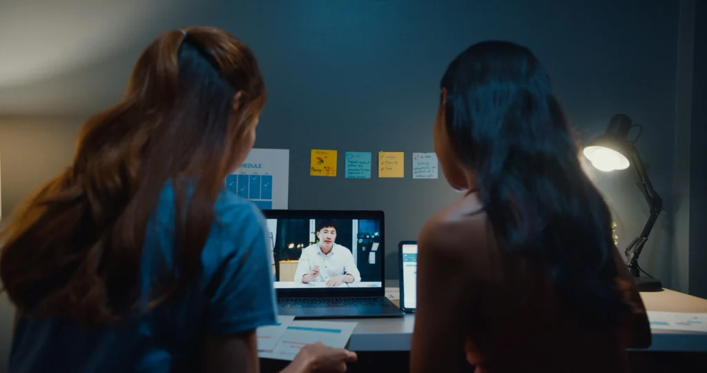 Two women sit at a desk watching a man speak on a laptop screen during a video call; notes and a lamp are visible on the desk and wall.