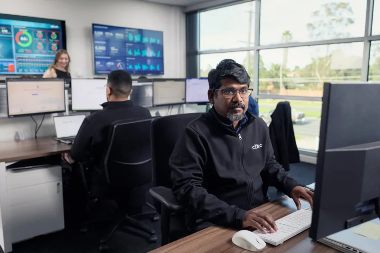 Three people work at computer stations in a modern office with multiple monitors displaying data and graphs; large windows provide natural light.