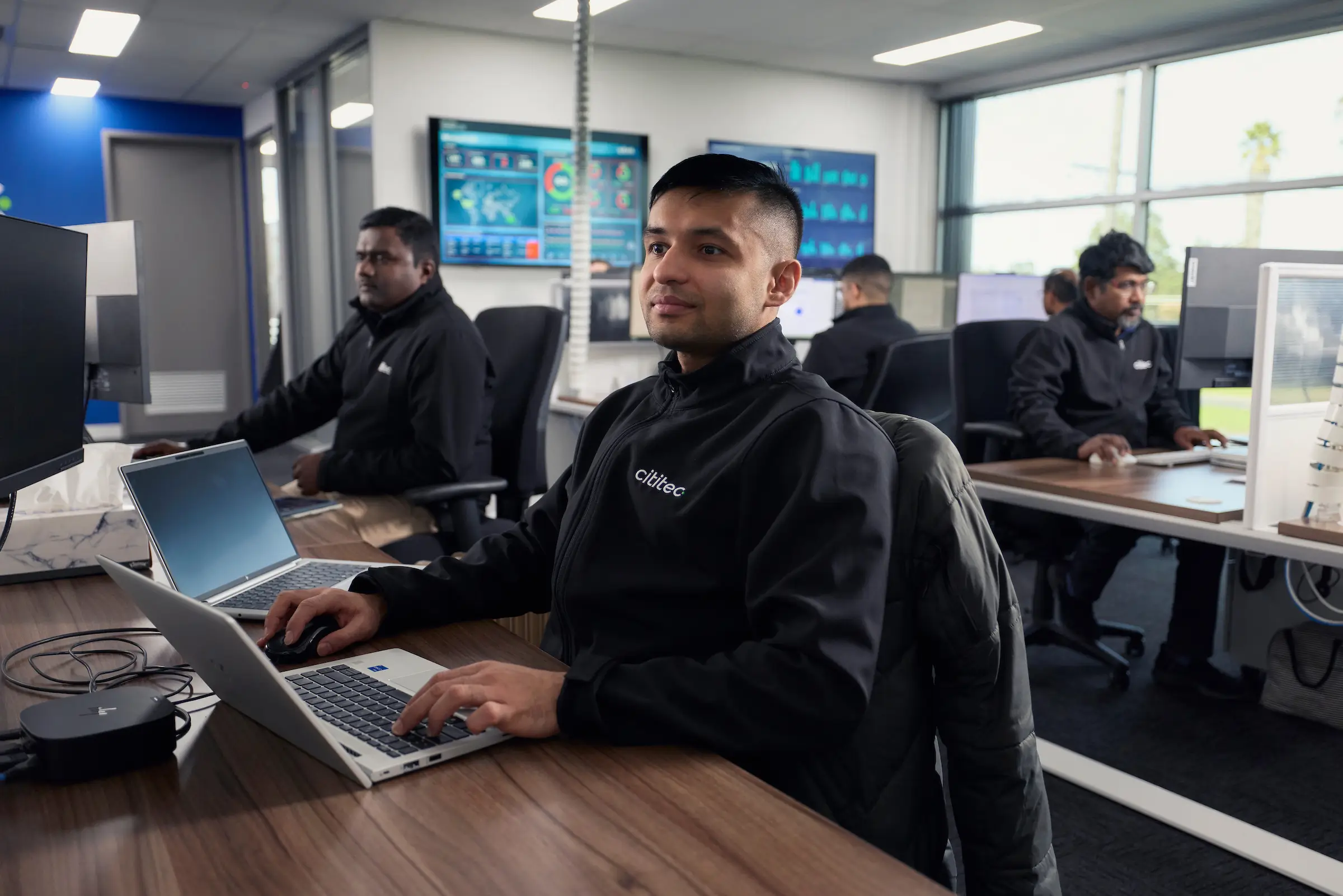 Several people in matching black jackets work on laptops and desktop computers in a modern office with large windows and wall displays.