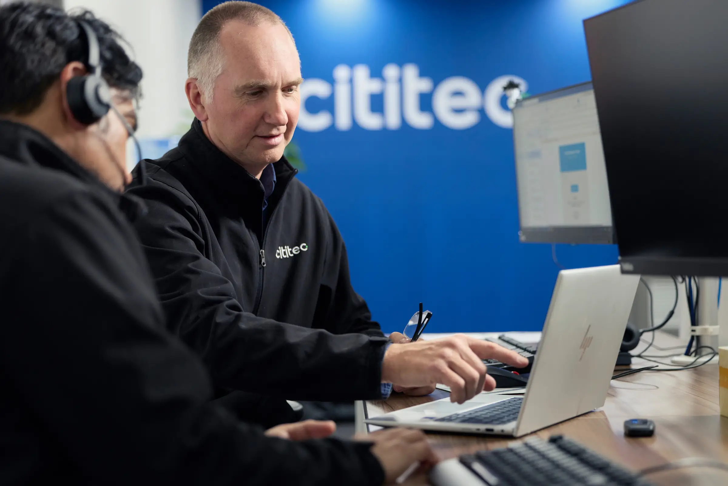 Two men working at laptops in an office, one pointing at a screen, with "cititec" logo visible in the background.